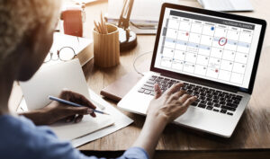 An image of a business woman sitting at her office desk, checking the monthly calendar on her laptop, with one of the dates circled in red.